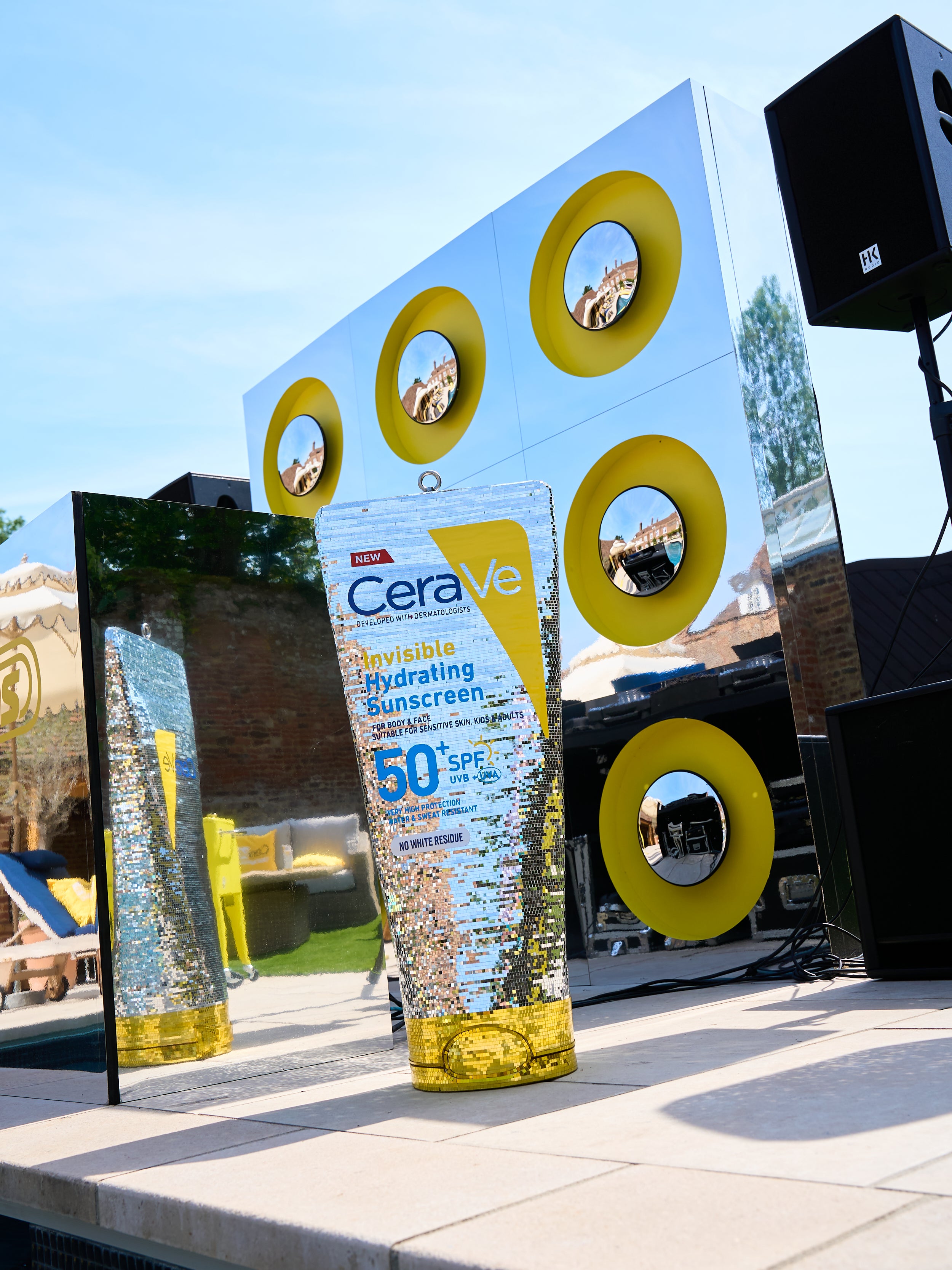 A disco ball CeraVe Invisible Hydrating Sunscreen bottle with a clear body and yellow cap stands outdoors on a patio in front of a reflective mirror and a large decorative wall with yellow circular cutouts. created by sophie lopez, town girl disco in london 