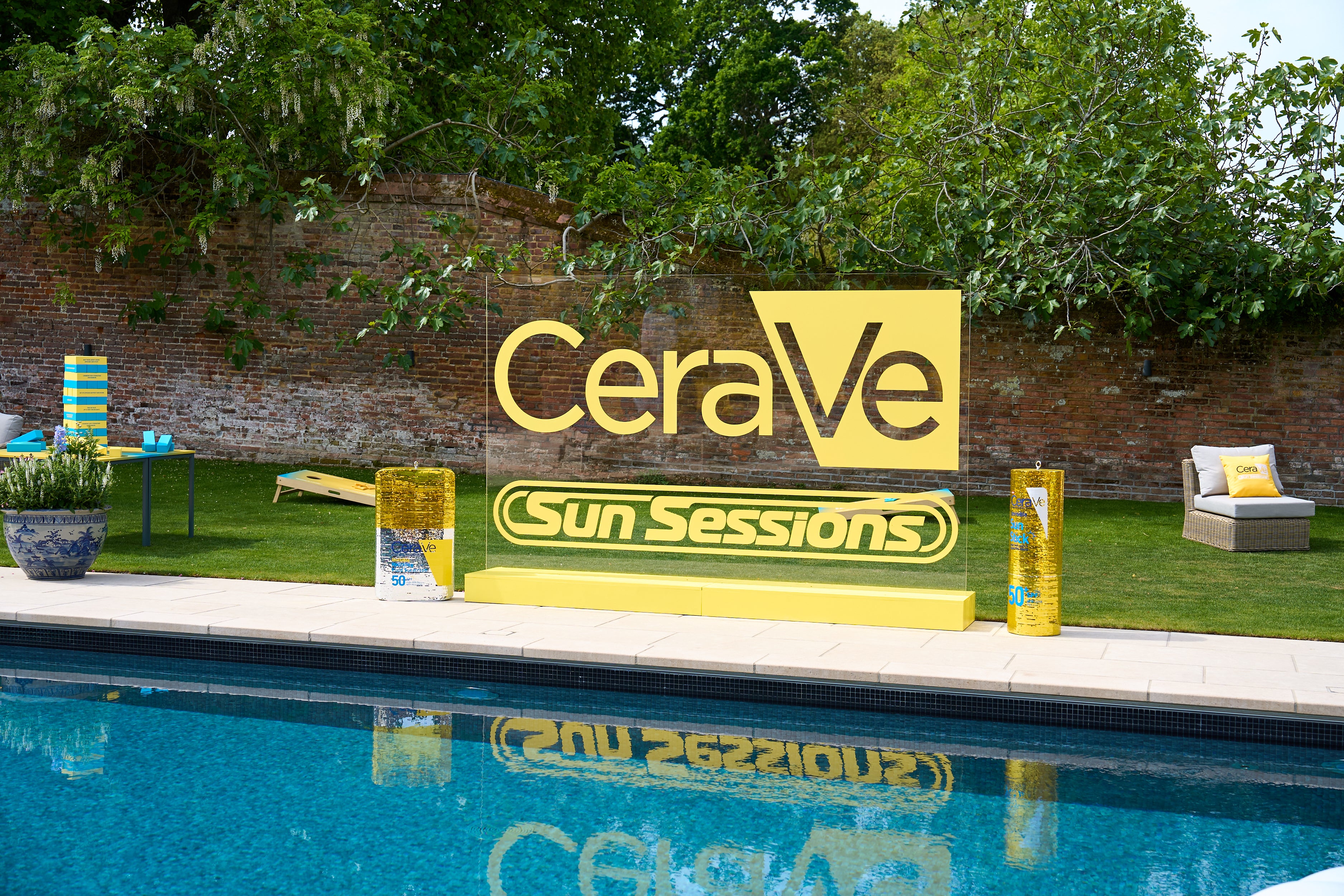 2x Large disco ball CeraVe sculptures made by London based disco ball artist, Sophie Lopez aka town girl disco beside a large yellow outdoor sign reading 'CeraVe Sun Sessions' stands beside a swimming pool,